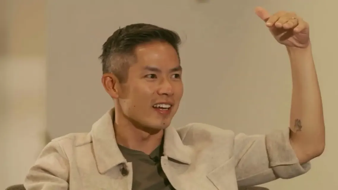 Brandon in the middle of a story, one hand up, in a beige jacket